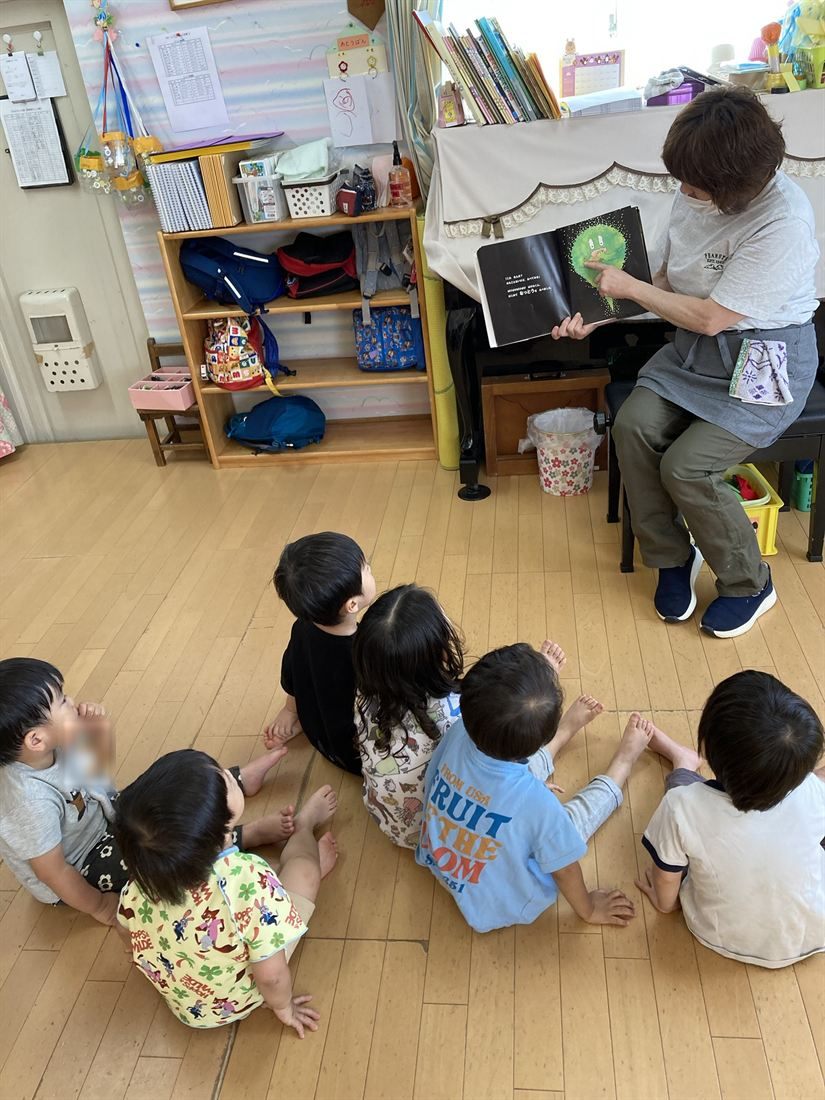先生が絵本を開き、子どもたちが床に座って見上げている