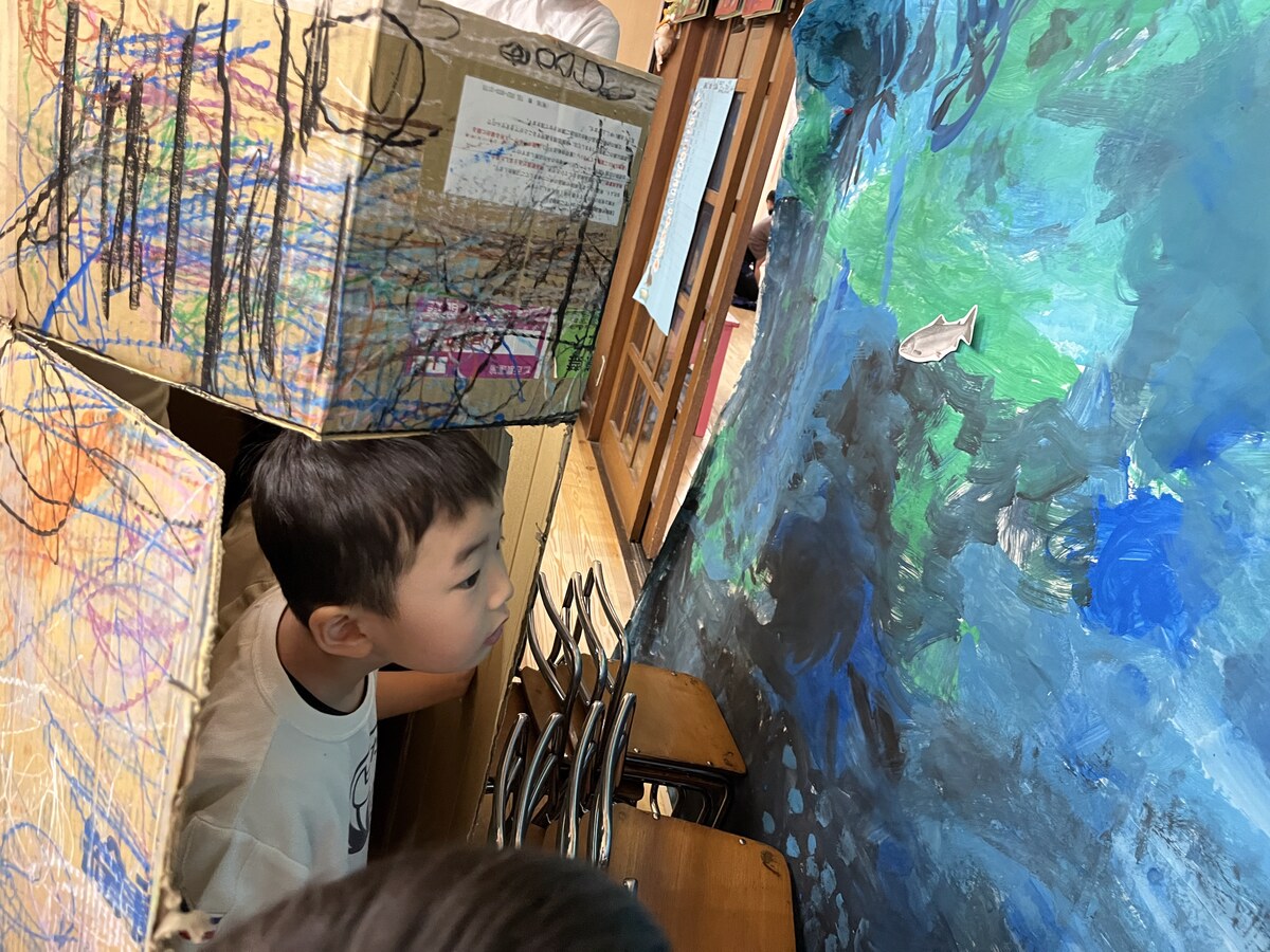 A child peeking from a cardboard submarine they built, looking at the ocean mural